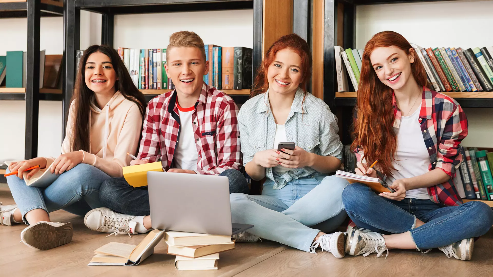 Group of smiling teenagers doing homework while sitting on a floor at the library with laptop computer and mobile phone